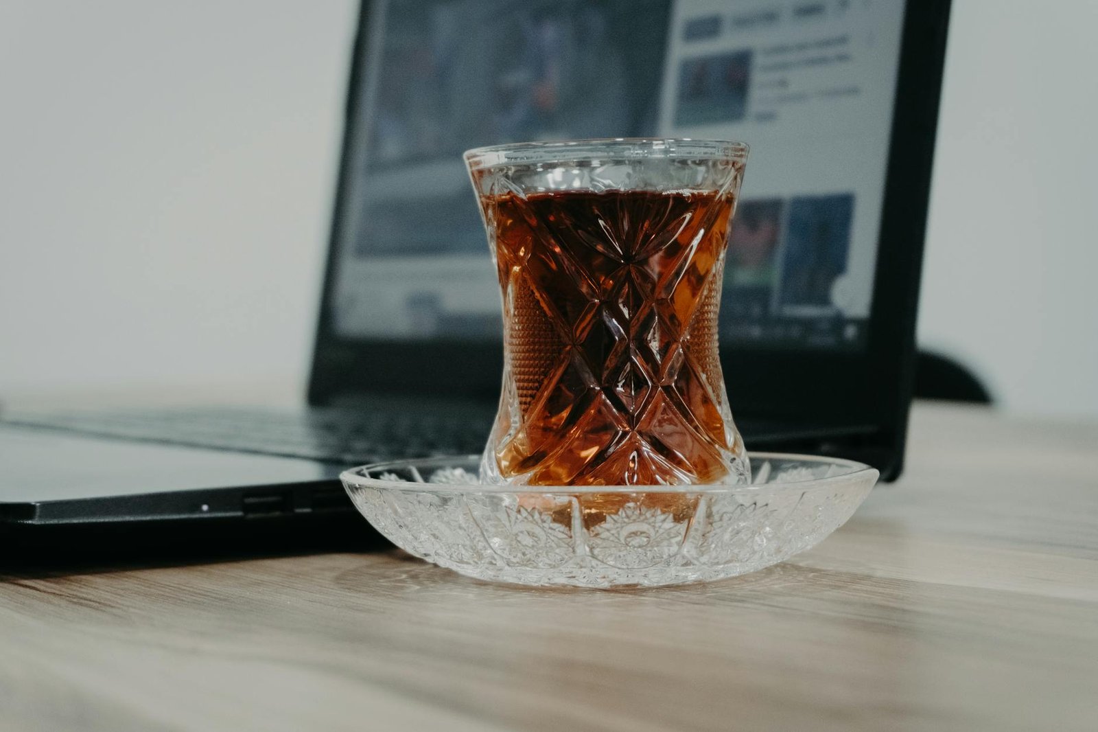 A clear glass of black tea sits on a wooden desk beside a laptop, catching warm light and showing a deep amber colour. The scene evokes a short work break or home office moment.