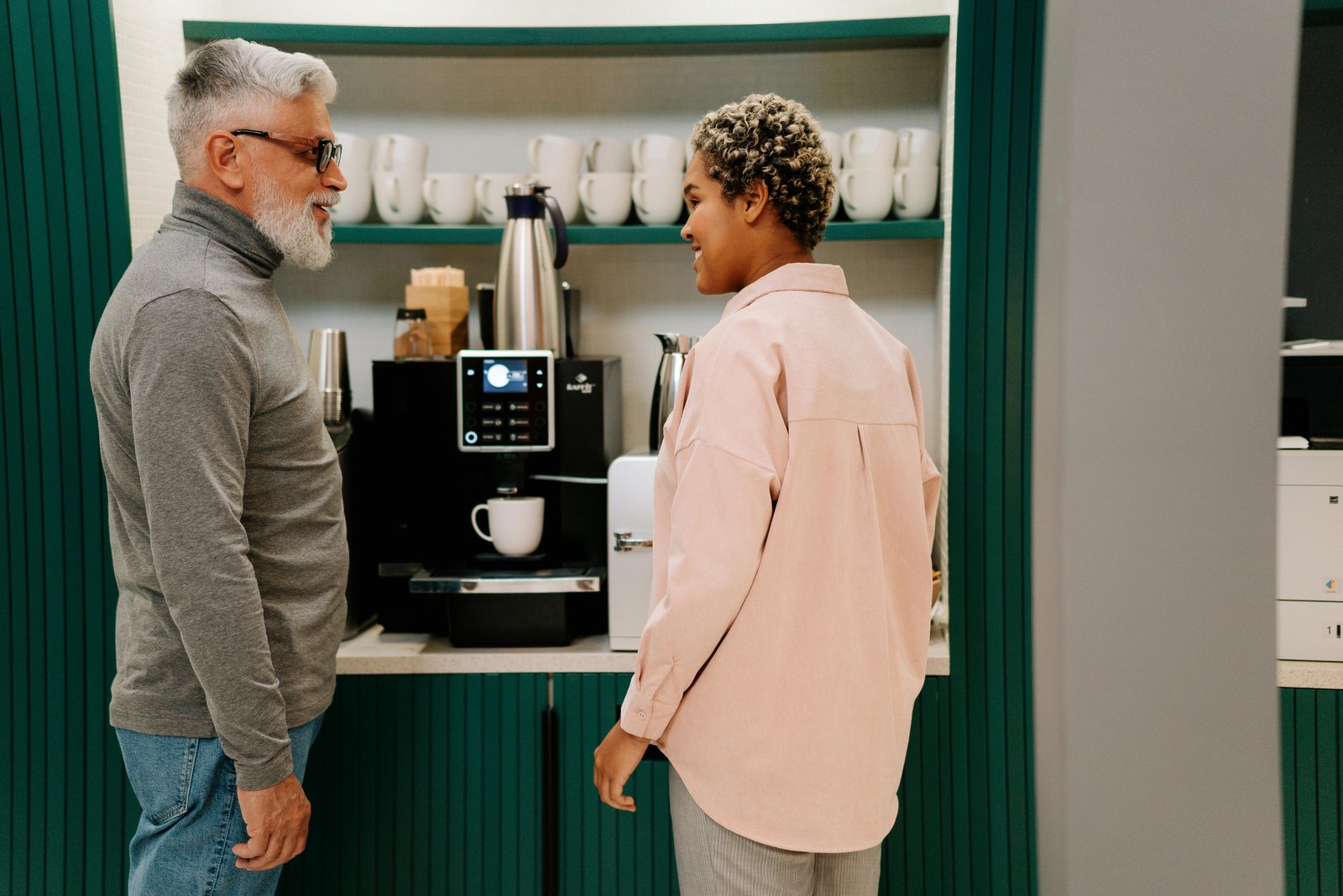 Two colleagues chat while preparing coffee at a modern office coffee machine in a bright break room.