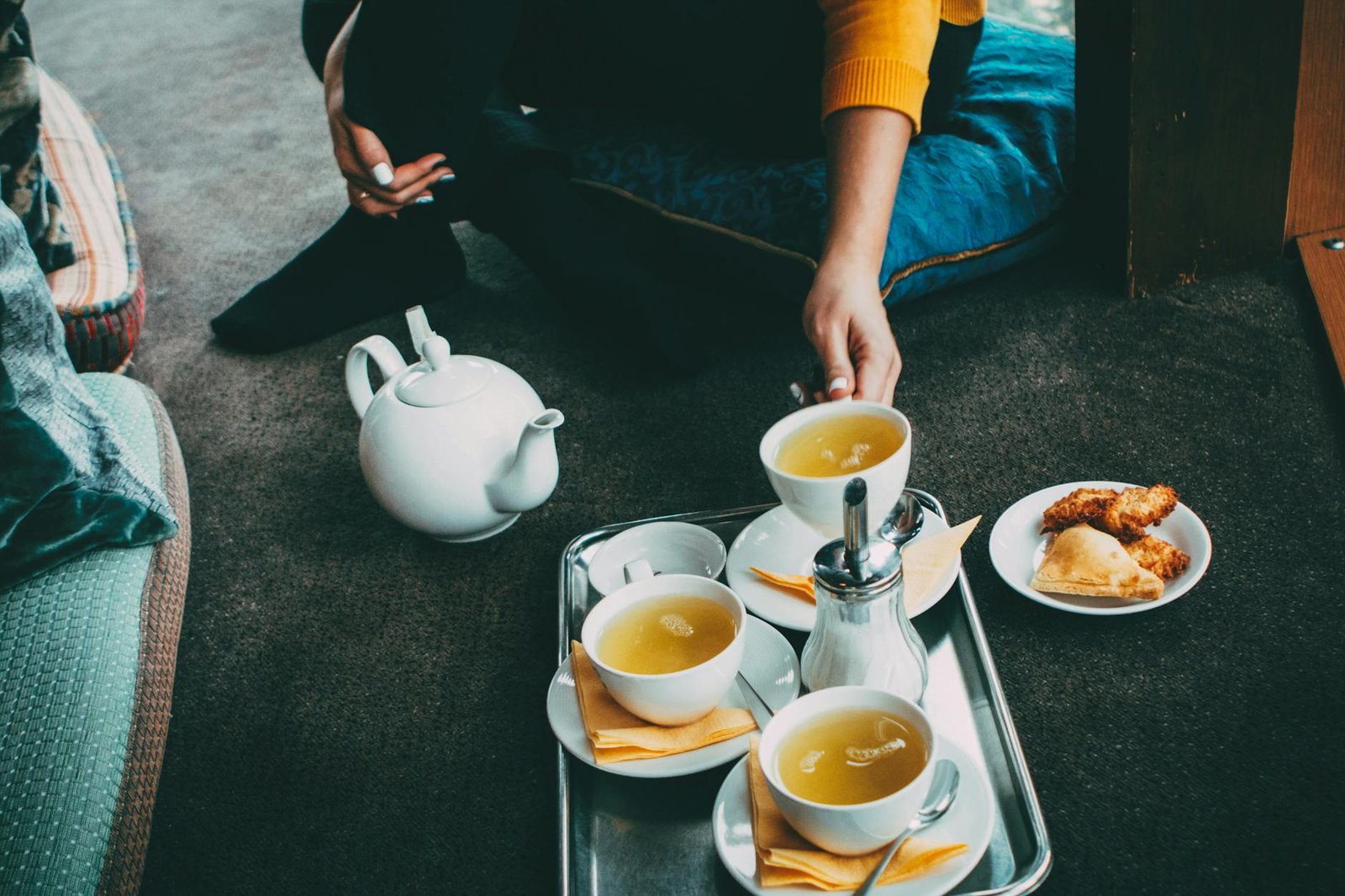 A person's hands holding a steaming cup of tea next to a glass teapot and small snacks on a wooden table, suggesting a calm tea break.