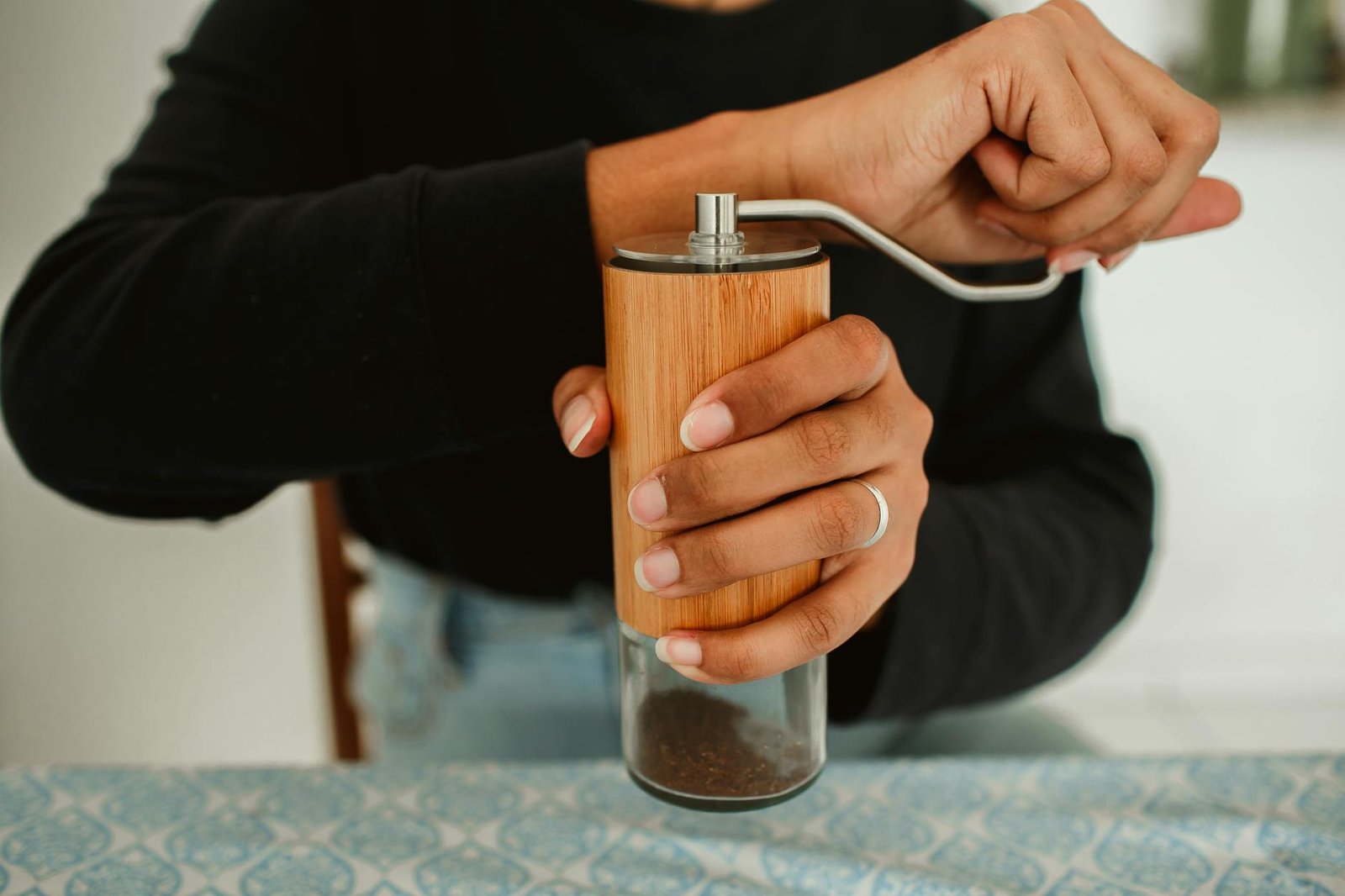 Person grinding coffee beans with a wooden manual burr grinder, hands turning the crank over a small jar of freshly ground coffee. Loose beans are visible on the counter.