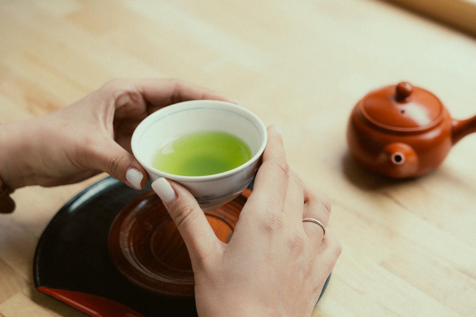 Hands cradling a steaming cup of green tea beside a clay teapot on a wooden surface, lit by soft natural light.